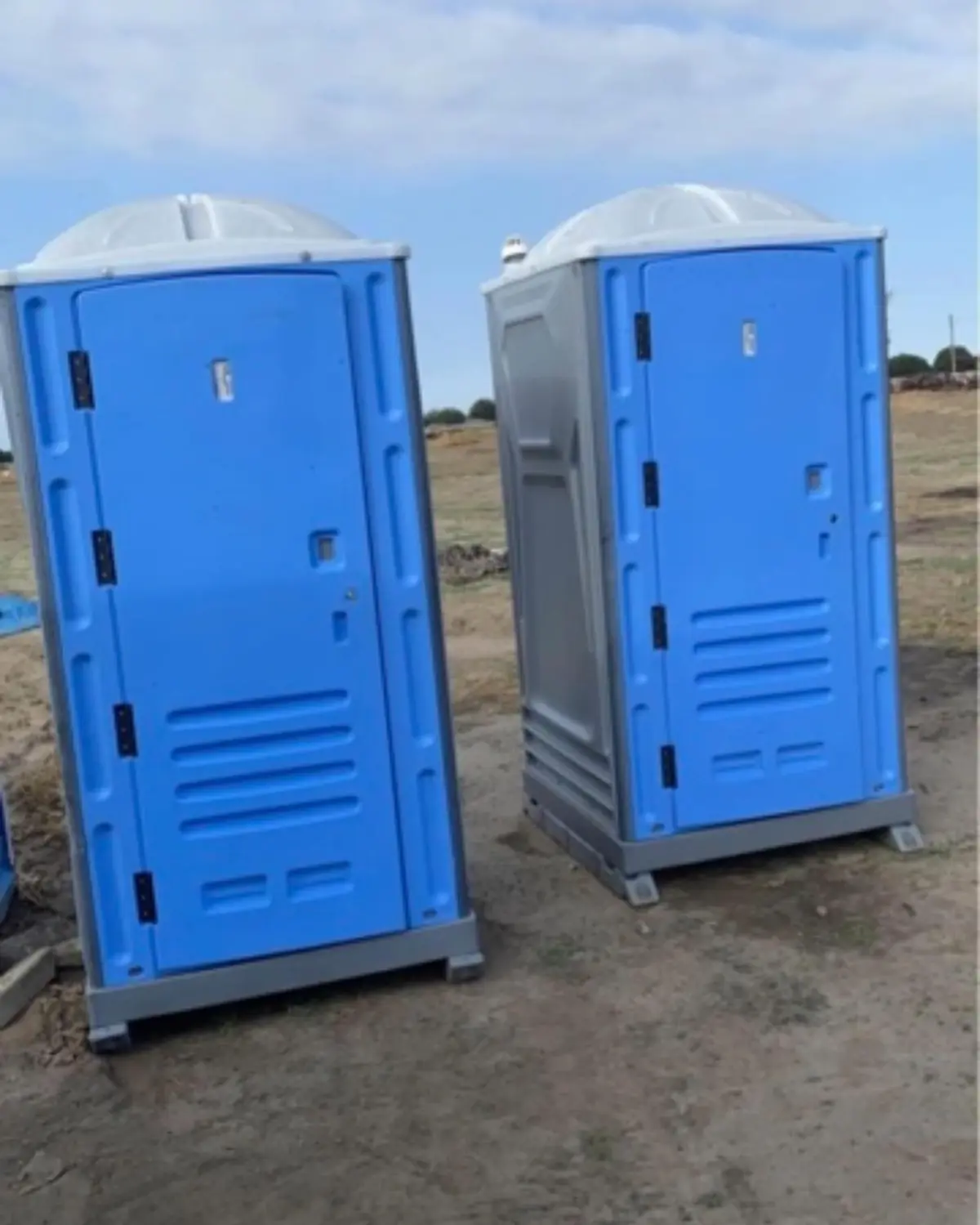 Two blue standard porta potties set on open dirt ground under a clear sky