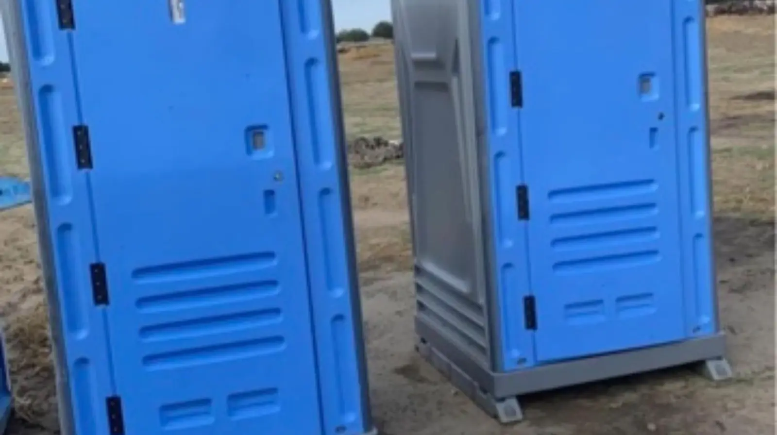 Two blue standard porta potties set on open dirt ground under a clear sky